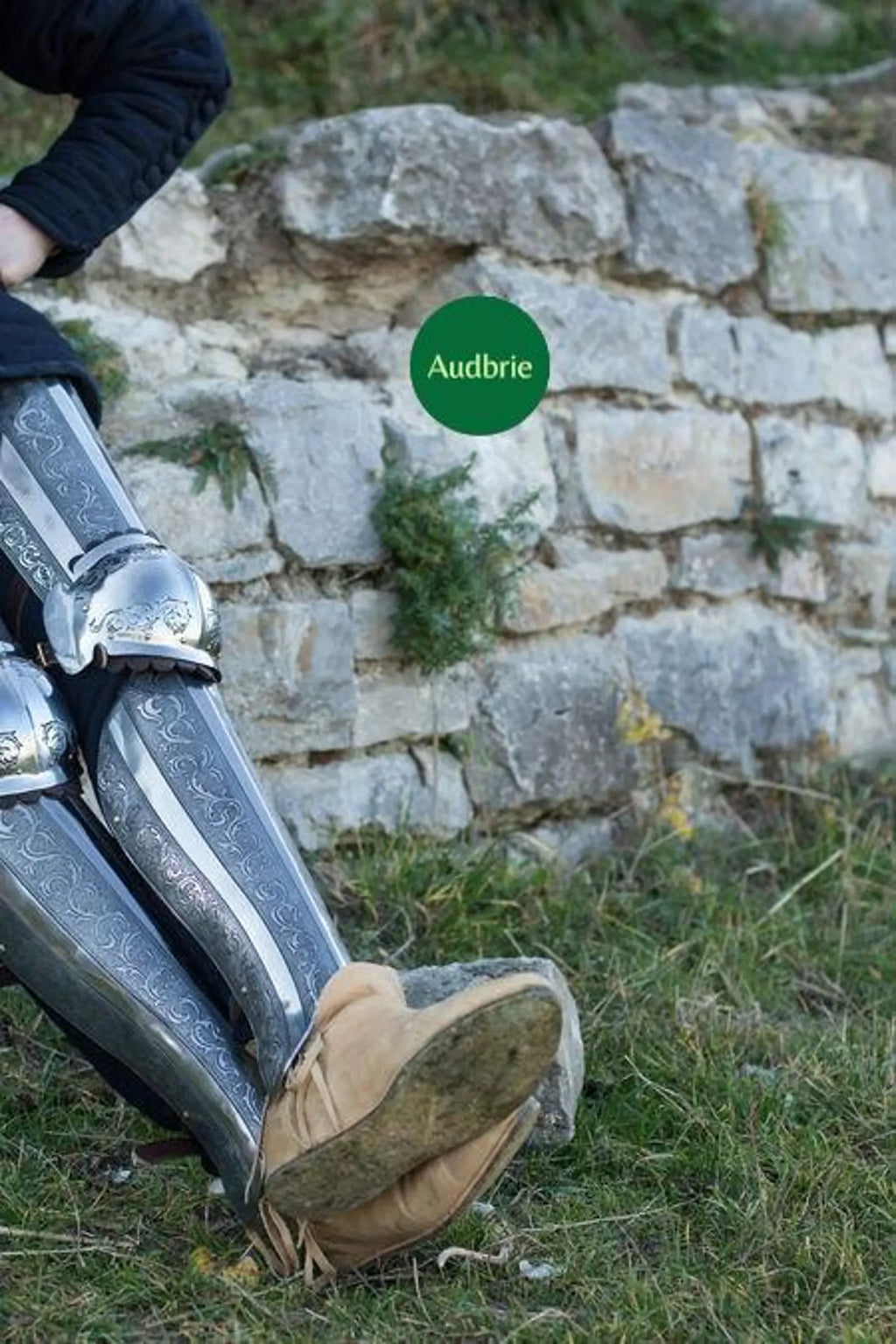 Person wearing metallic knee pads and brown shoes standing in front of a stone wall with 'Audrie' branding.