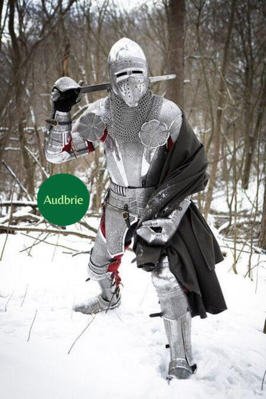 Person wearing a full medieval knight armor suit with steel helmet, chainmail, plate armor, and sword while standing in a snowy forest.
