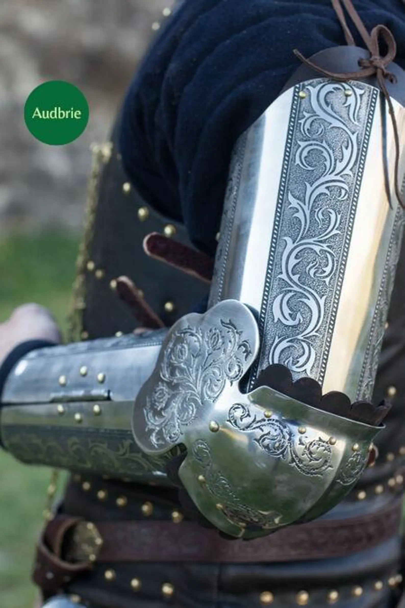 Decorative metal arm guard with intricate patterns, worn by a person, against a natural background.