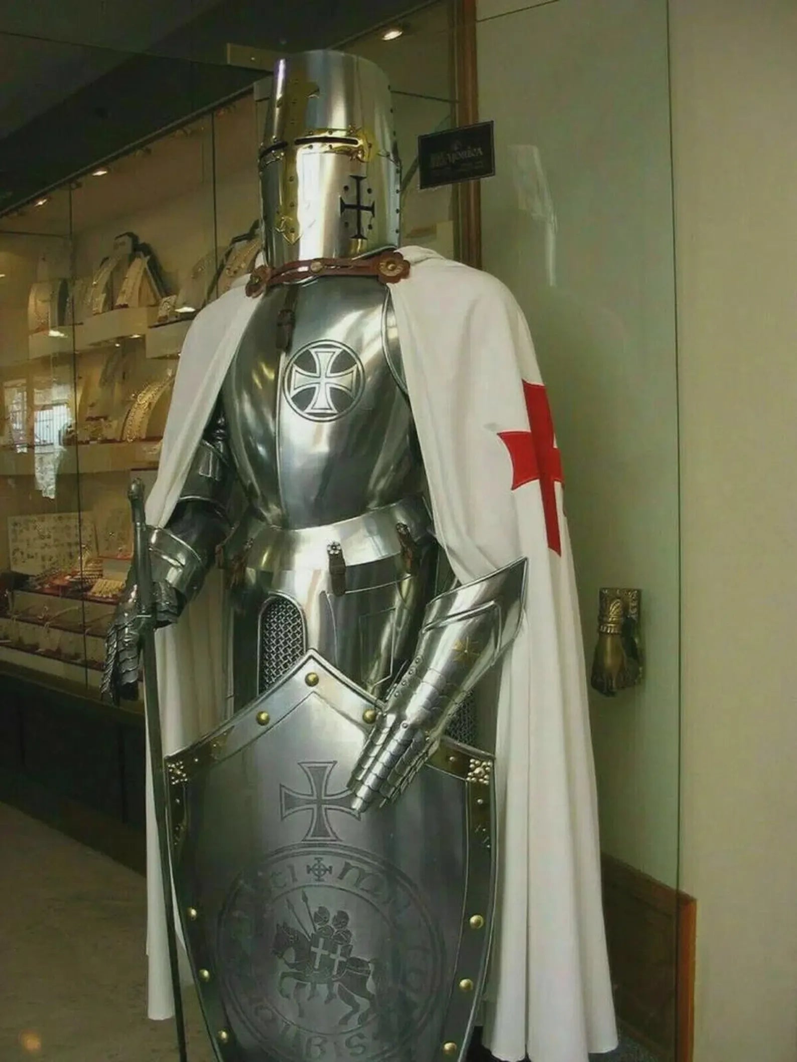 Medieval knight's armor with a white cape and red cross, displayed indoors.